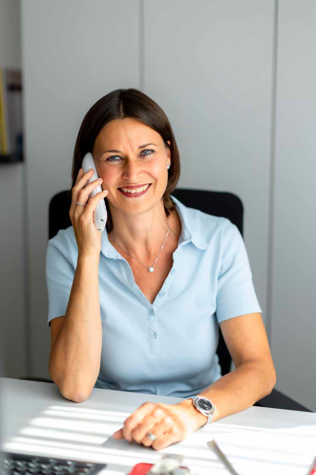 Eine Frau mit dunkelbraunem Haar telefoniert lächelnd in einem Büro. Sie trägt ein hellblaues Blusen-Shirt.