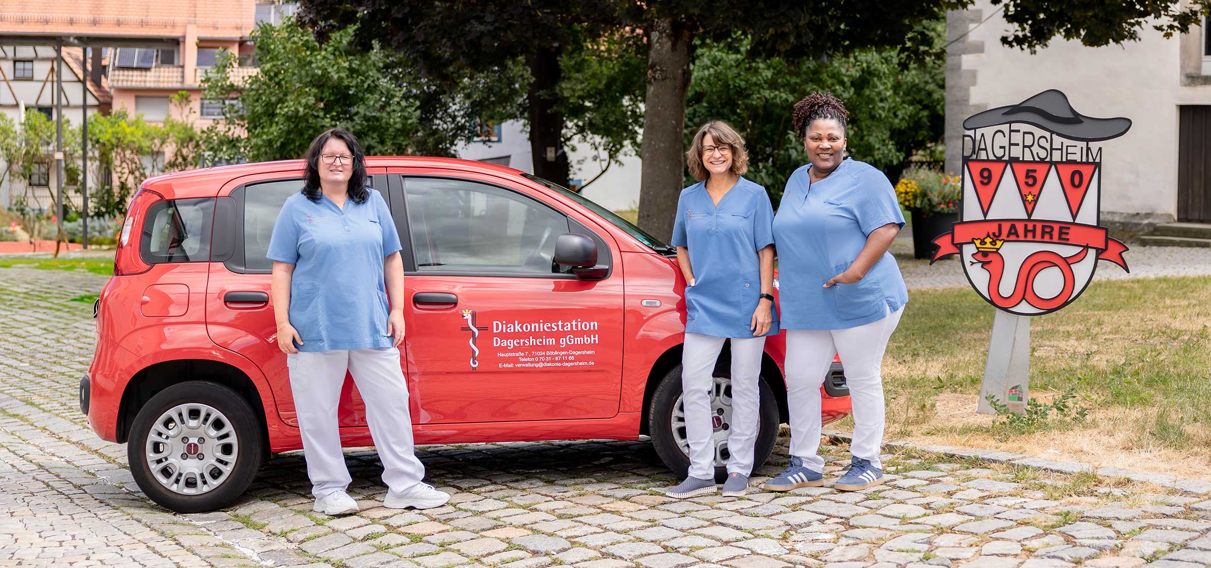 Drei Mitarbeiterinnen der Diakoniestation Dagersheim stehen in blau-weißer Berufskleidung vor einem roten Dienstfahrzeug auf einem gepflasterten Platz neben einem Schild mit der Aufschrift „Dagersheim 950 Jahre“.