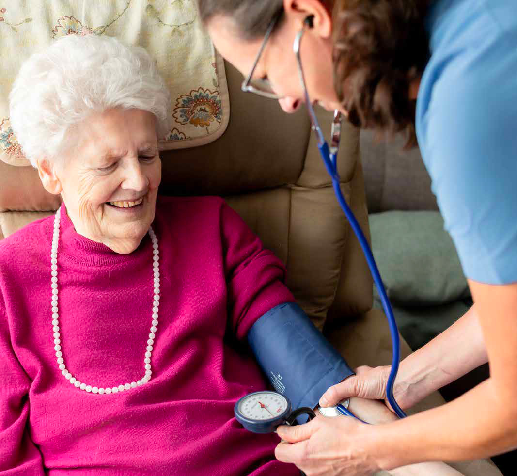 Eine ältere Frau in pinkem Pullover lächelt während einer Blutdruckmessung durch eine Pflegefachperson mit Stethoskop. Die Pflegefachperson trägt blaue Berufskleidung.